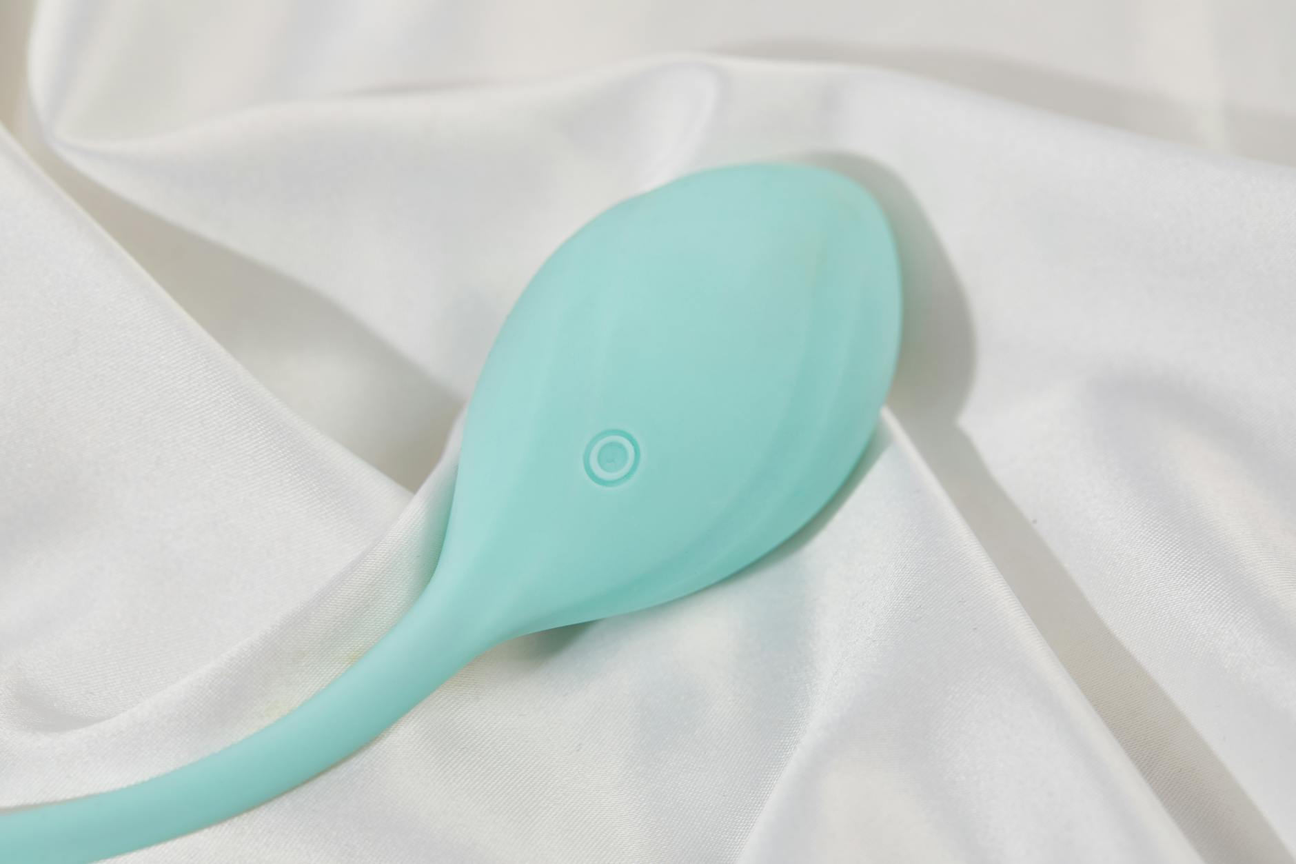 A teal clitoral vibrator on soft white silk fabric, representing gentle pleasure during recovery