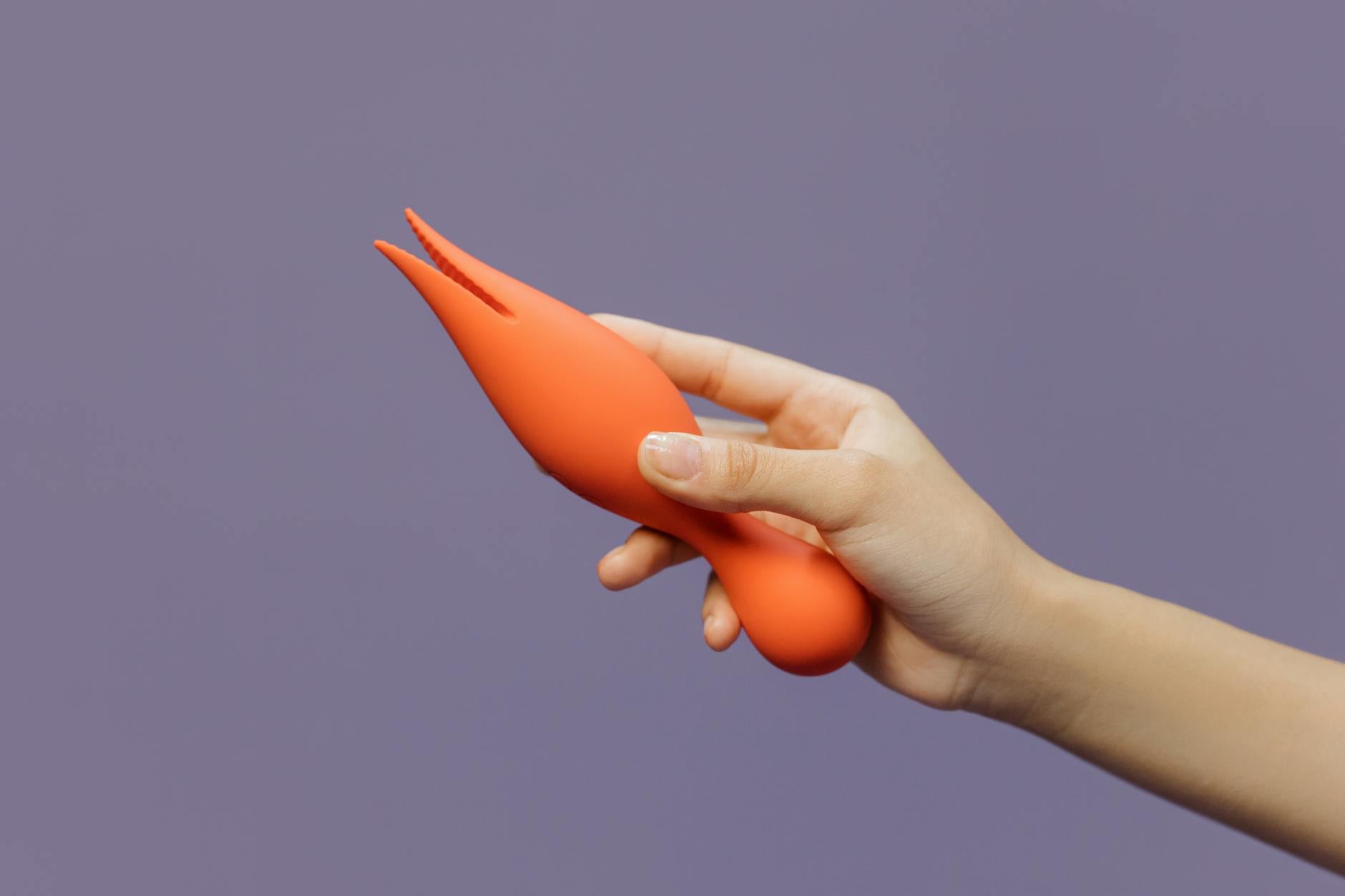 A hand holding a blue silicone clitoral vibrator against a purple background, representing self-directed pleasure and healing.